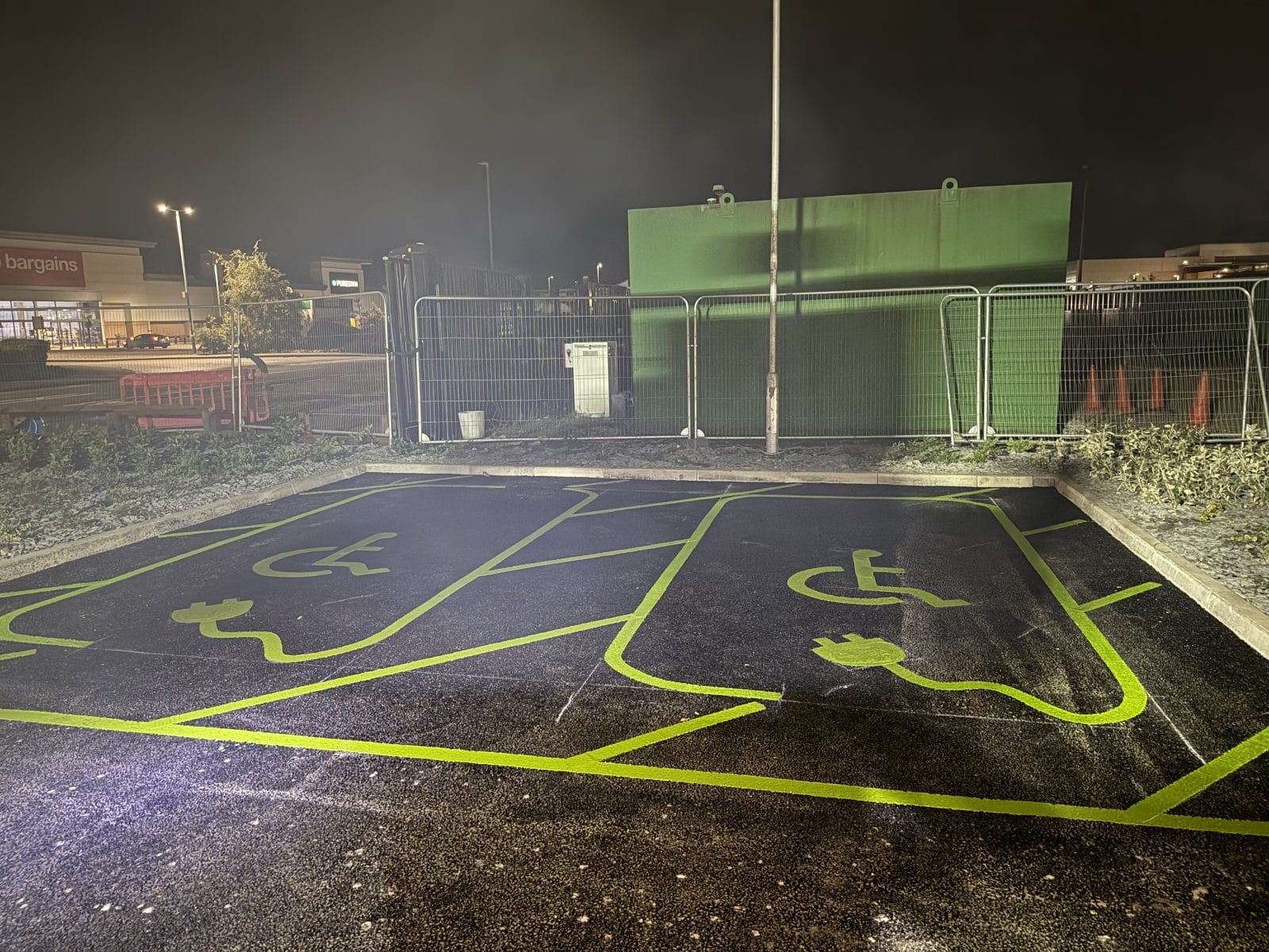 Nighttime view of a parking area with bright yellow road markings and a green building in the background.