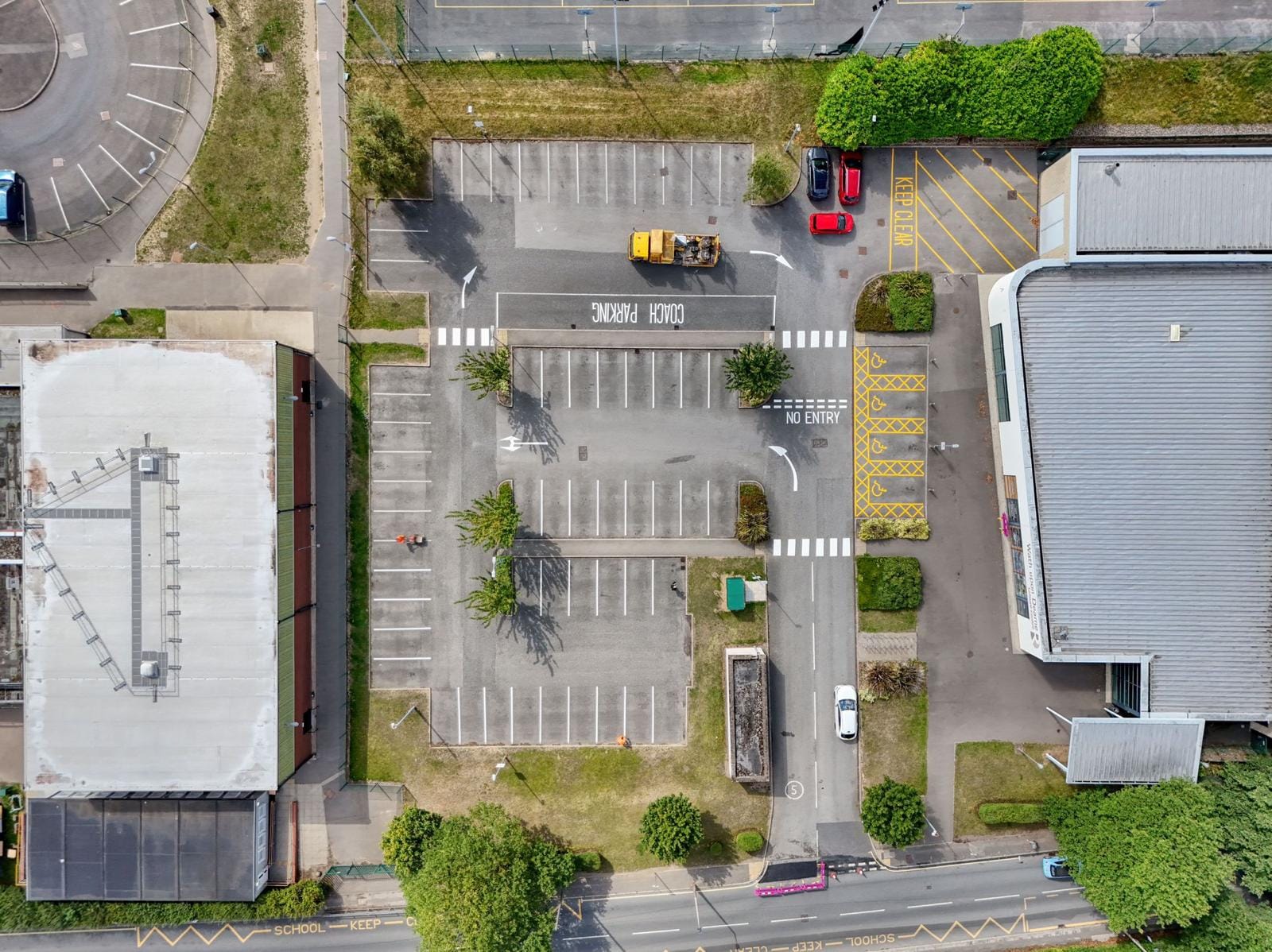 Aerial view of a large parking lot with clearly marked white parking spaces, yellow no-parking zones, handicap spots, directional arrows, and a coach parking area. Surrounding buildings, greenery, and a few parked vehicles are visible.
